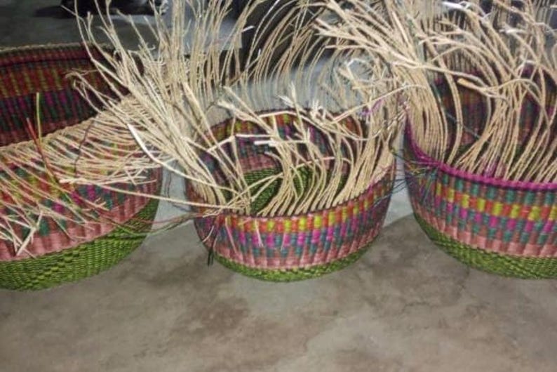 May include: Three round woven baskets featuring a vibrant multicoloured checkered pattern in shades of green, pink, and yellow. Each basket has long, unfinished natural tan fibres extending upwards from the top rim, showing the construction process.