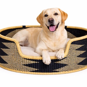 May include: A golden Labrador lies in a black and gold woven dog bed. The bed has a rounded shape with a geometric pattern. The dog is smiling with its tongue out.