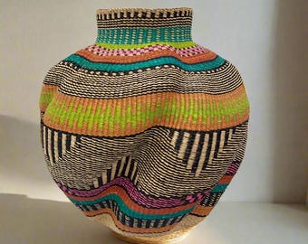 Könnte beinhalten: Ein runder, bauchiger Korb aus geflochtenem Material. Der Korb besteht aus Naturfasern und hat ein farbenfrohes geometrisches Muster in den Farben Schwarz, Weiß, Grün, Orange, Rosa und Türkis.