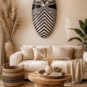Puede incluir: Un salón con un sofá blanco, cojines beige, una mesa de centro redonda de madera y una cesta tejida. Sobre el sofá, un gran elemento decorativo de pared con un patrón en blanco y negro. Jarrones y una alfombra completan la cálida decoración en tonos neutros.