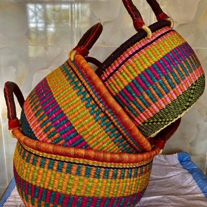 May include: Two round woven baskets featuring vibrant horizontal stripes in shades of pink, orange, yellow, teal, and dark green. Each basket has two sturdy dark brown leather-wrapped handles attached to the rim for easy carrying and transport.