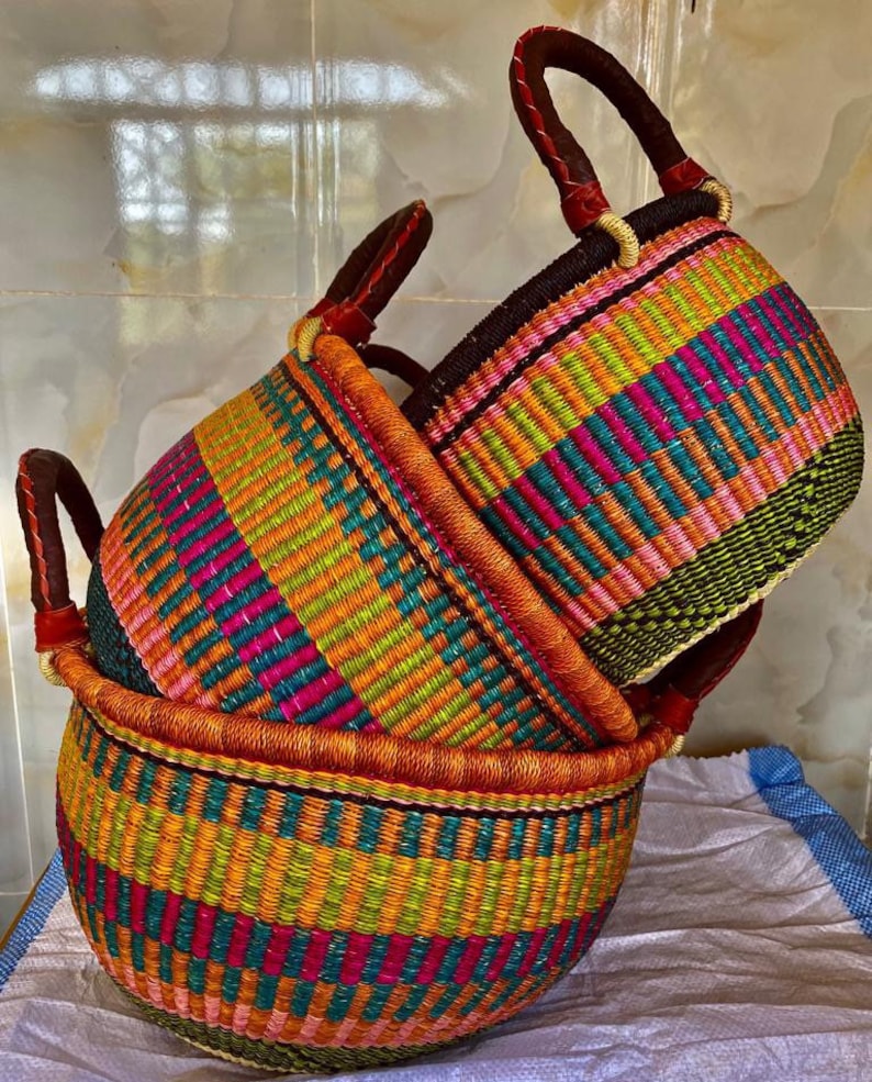 May include: Two round woven baskets featuring vibrant horizontal stripes in shades of pink, orange, yellow, teal, and dark green. Each basket has two sturdy dark brown leather-wrapped handles attached to the rim for easy carrying and transport.