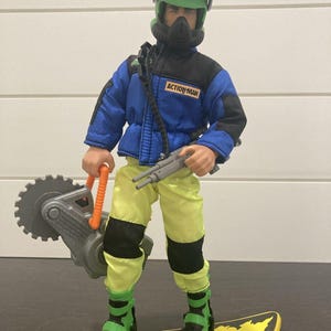 May include: An Action Man figure wearing a blue and black jacket with a green helmet and visor, yellow pants with black knee patches, and green boots. The figure holds a gray circular saw and a gray rifle while standing on a yellow snowboard.