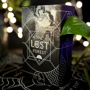May include: A black and white tarot deck box with the text "The Lost Forest Tarot" on the front. The box is sitting on a black surface with a white design.