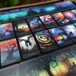 Op de afbeelding: Een set tarotkaarten met kleurrijke, abstracte kunstwerken. Elke kaart heeft een uniek ontwerp met een titel onderaan. De kaarten liggen op een zwart oppervlak.