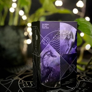 Op de afbeelding: De doos van het The Lost Forest Tarot Deck heeft een paars en zwart ontwerp met een draak en een vrouw met vleugels. De doos is gelabeld "3rd Edition 80 Card Tarot Deck".