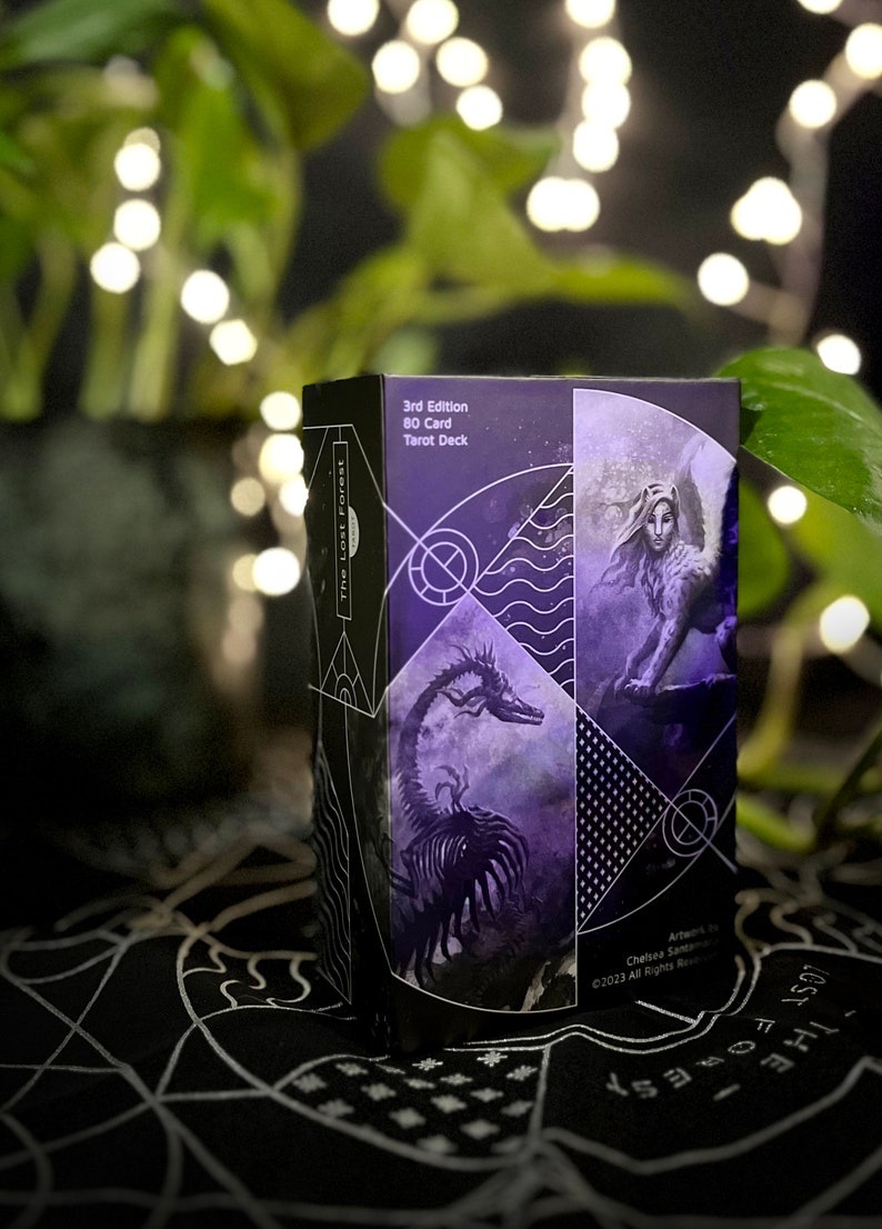 Op de afbeelding: De doos van het The Lost Forest Tarot Deck heeft een paars en zwart ontwerp met een draak en een vrouw met vleugels. De doos is gelabeld "3rd Edition 80 Card Tarot Deck".