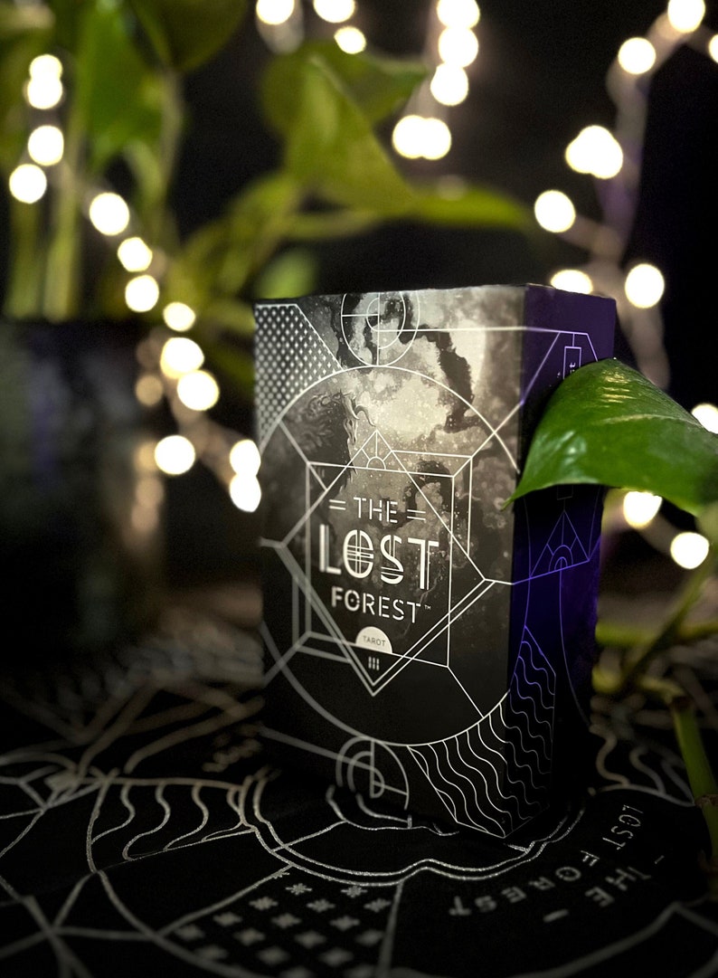 Op de afbeelding: Een zwarte en witte tarotdeckdoos met de tekst "The Lost Forest Tarot" op de voorkant. De doos staat op een zwart oppervlak met een wit ontwerp.