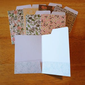 May include: A collection of decorative paper folders with floral and patterned designs. The folders feature various colors, including pink, green, and yellow, and are arranged on a wooden surface. One folder is open, revealing a light blue floral pattern.