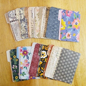 May include: A collection of decorative file folder dividers with various patterns and colors. The dividers feature floral, botanical, and vintage-inspired designs, including text and patterned backgrounds. The folders are arranged on a wooden surface.