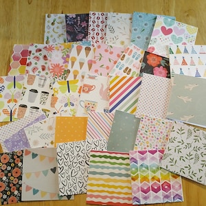May include: A collection of patterned paper squares in various designs, including floral, hearts, stripes, and geometric patterns. The color palette includes pink, blue, green, and yellow. The papers are arranged on a wooden surface.
