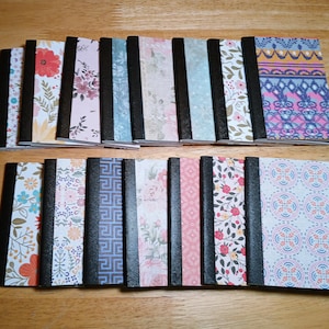 May include: A collection of sixteen small rectangular notebooks arranged in two rows. Each notebook features a black spine and a unique patterned cover, including floral, geometric, and abstract designs in various colors like pink, blue, and orange.