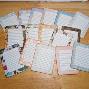 May include: A collection of lined note cards with decorative borders and floral or butterfly patterns. The cards have a rectangular shape with rounded corners and are arranged on a wooden surface.