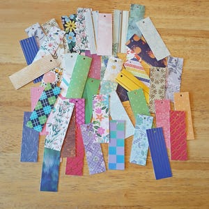 May include: A collection of colorful paper bookmarks with various patterns, including floral, plaid, and geometric designs. Each bookmark has a small hole at the top. The bookmarks are scattered on a wooden surface.