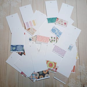 May include: A collection of white rectangular tags with decorative patterned paper accents at the bottom. The tags feature various designs, including floral, geometric, and striped patterns. Each tag has a small hole at the top.