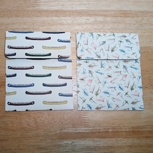 May include: Two small, square paper envelopes. One features a pattern of canoes in blue, brown, and yellow on a white background. The other has a pattern of fishing lures in blue, pink, and green on a white background. Both have a flap closure.