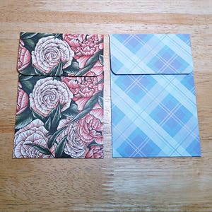 May include: Two rectangular paper envelopes. The left envelope features a detailed floral pattern with pink roses and dark green leaves on a black background. The right envelope displays a light blue and white diagonal plaid pattern.