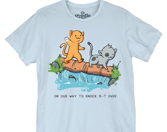 May include: A light blue t-shirt featuring a graphic of an orange cat and a gray spotted cat walking across a log over a blue stream. Below the illustration, the text reads: ON OUR WAY TO KNOCK S--T OVER. The shirt has short sleeves and a crew neck.