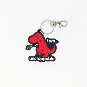 May include: A red dinosaur keychain with the words "I am... unstoppable". The dinosaur is holding a selfie stick. The keychain has a silver clasp and a keyring.