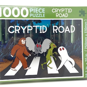 Op de afbeelding: Een 1000-delige puzzel met een cartoonillustratie van Bigfoot, Mothman, Lizardman en een geest die een weg oversteken. De puzzeldoos is groen en heeft de tekst "CRYPTID ROAD" erop. De puzzel is 750 mm bij 510 mm.