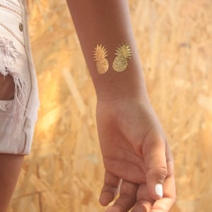 May include: Two gold pineapple temporary tattoos are displayed on a person's wrist. One is solid gold, and the other has a glittery effect. The person is wearing light-colored shorts. The background is a light brown wood.