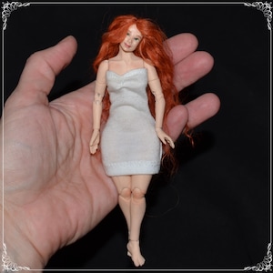 Puede incluir: Una pequeña muñeca con el pelo largo y rizado rojo que lleva un vestido blanco sin tirantes. La muñeca está posada con los brazos ligeramente levantados y las piernas ligeramente dobladas.