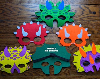 Può includere: Una collezione di maschere di dinosauro in feltro colorato in verde, rosso, arancione, giallo e viola. Ogni maschera presenta un design unico con corna e fori per gli occhi. Una maschera ha il testo "Zander's 3rd Birthday".