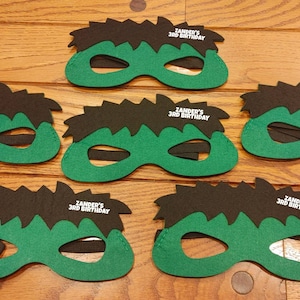 May include: Green and black felt masks with the text "Zander's 3rd Birthday" printed in white. The masks have a jagged, spiky design at the top and an elastic band for wearing. The masks are arranged on a wooden surface.