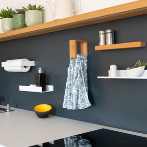 May include: A modern kitchen with white cabinets and a black countertop. A black electric stovetop is installed in the countertop. A stainless steel sink is installed in the countertop. A white paper towel holder is mounted on the wall. A white shelf with a small bowl is mounted on the wall. A wooden shelf with a tea kettle and plants is mounted on the wall. A towel is hanging on a hook on the wall.