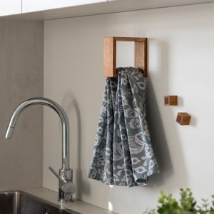 May include: A wooden towel rack with a square design, mounted on a white wall. A patterned kitchen towel hangs from the rack. Two small wooden blocks are attached to the wall.