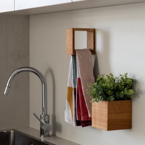 May include: A wooden wall-mounted shelf with a square opening holds a stack of colorful kitchen towels. A second wooden shelf with a square opening holds a small potted plant with green leaves. The shelves are mounted on a white wall above a stainless steel kitchen sink.