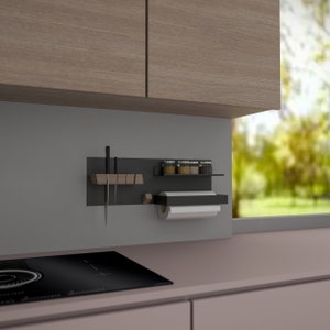 Pode incluir: Um organizador de cozinha de metal preto com um suporte magnético para facas, um suporte para papel toalha e uma pequena prateleira para guardar especiarias. O organizador está montado em uma parede cinza.