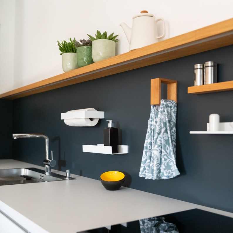 May include: A white kitchen with a black wall and a white countertop. There are several shelves with plants, a towel rack, a soap dispenser, and a paper towel holder. There is a sink and a stovetop in the image.