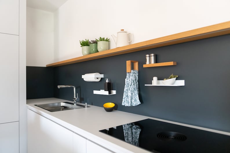 May include: A modern kitchen with white cabinets and a black countertop. A black electric stovetop is installed in the countertop. A stainless steel sink is installed in the countertop. A white paper towel holder is mounted on the wall. A white shelf with a small bowl is mounted on the wall. A wooden shelf with a tea kettle and plants is mounted on the wall. A towel is hanging on a hook on the wall.