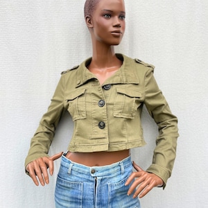 Puede incluir: Chaqueta corta verde oliva con cuatro bolsillos delanteros y botones negros. La chaqueta se combina con vaqueros de mezclilla azul claro. El conjunto se exhibe en un maniquí sobre un fondo blanco.