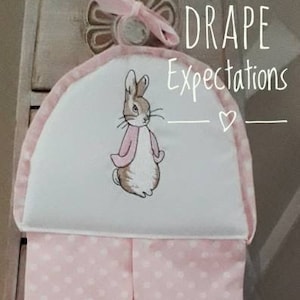 May include: A pink and white nappy stacker with a white bunny rabbit illustration. The stacker has a pink polka dot pattern and says "DRAPE Expectations" in black text.