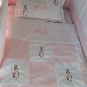 May include: A pink and white baby crib bedding set with a patchwork design featuring embroidered Peter Rabbit characters. The set includes a pillow and a quilt with the name "Isla" embroidered on both.