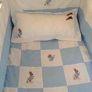 May include: A blue and white checked cot bedding set with a Peter Rabbit theme. The bedding features a white pillow with two red carrots embroidered on it and a blue and white checked quilt with Peter Rabbit embroidered on it.