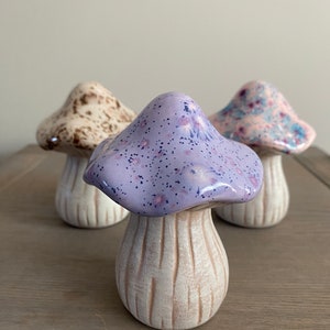 May include: Three ceramic mushroom figurines. The mushroom on the left is brown and white with a speckled pattern. The mushroom in the center is purple with a white base and a speckled pattern. The mushroom on the right is pink and white with a speckled pattern.