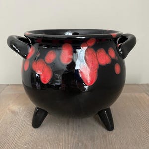 May include: A glossy black ceramic cauldron-style pot featuring two side handles and three short legs. The exterior surface is decorated with irregular, vibrant red spots that appear to drip down from the rim, creating a high-contrast pattern.