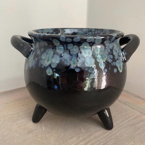 May include: A black ceramic cauldron with three legs and two handles. The rim and upper portion of the cauldron feature a speckled blue and green glaze. The cauldron has a glossy finish and sits on a wooden surface.