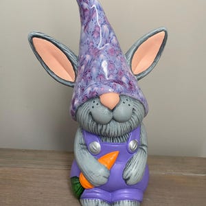 May include: A ceramic gnome-bunny figurine with a purple hat and overalls. The hat has a speckled design, and the ears are gray with pink insides. The figurine is holding a carrot and has a smiling face.