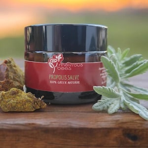 Puede incluir: Un pequeño frasco de bálsamo de propóleo, etiquetado como "Melirious Bees Propolis Salve 100% Greek & Natural", se encuentra sobre una superficie de madera con follaje verde y material vegetal seco.