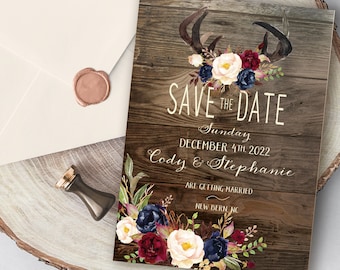 May include: A rustic "Save the Date" card with a wooden background, adorned with watercolor antlers and floral arrangements in burgundy, navy, and white. The card includes the date, names, and location of the wedding. An envelope with a wax seal is also present.