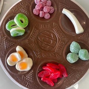 May include: A brown, circular Passover Seder plate filled with various candies. The candies include green ring-shaped gummies, pink round candies, white curved candies, green square candies, red fish-shaped candies, and egg-shaped candies.