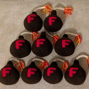 May include: A collection of nine handmade, dark brown knitted ornaments, each featuring a large red "F" and a white string with colorful yarn tassels. The ornaments are arranged on a light brown surface.