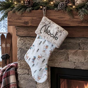 May include: A white Christmas stocking featuring a repeating pattern of illustrated reindeer and pine trees. The stocking has a plush, grey faux fur cuff embroidered with the name Nicole in dark thread and includes a metal hanging clip.