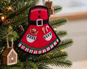 May include: A red felt apron ornament featuring a black belt with a silver buckle, two white pockets with green trim, and embroidered details including a gingerbread person, a wooden spoon, and a candy cane, hanging from a black ribbon.