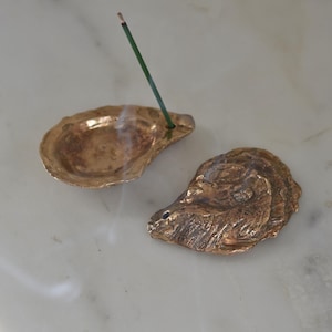 May include: Two bronze-colored oyster shell incense holders. One shell holds a green incense stick that is lit and emitting smoke. The other shell is empty. The shells are on a white marble surface.