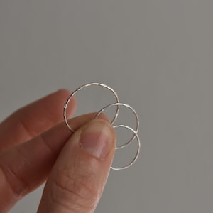 May include: A hand holds four small, delicate silver rings. The rings are circular with a textured surface. The rings are different sizes, with the largest ring at the top. The background is a solid gray.